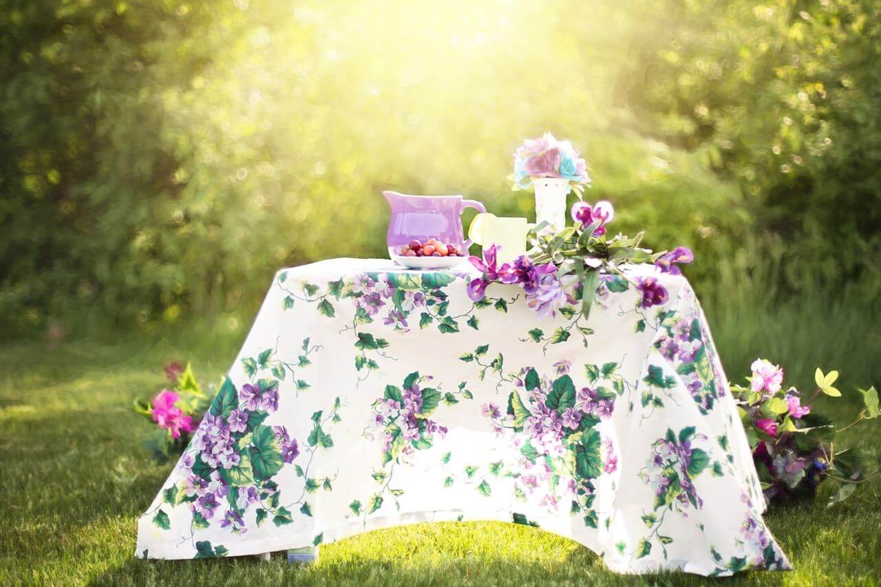 garden table covered with a very flowery tablecloth with a glass of lemonade, a pitcher and a bouquet of flowers on top.